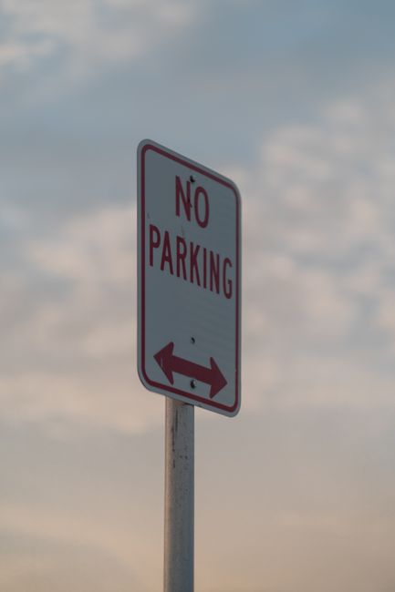 A vertical rectangular white 'No Parking' sign with red and black lettering, mounted on a metal pole situated on a street pavement under a partly cloudy sky. In the background, there are no vehicles or people visible. The sign indicates parking restrictions in the area around Brunswick Park, which is relevant for home relocation and furniture transport planning. The image may be used in the context of moving services by Man with Van Brunswick Park, highlighting loading rules and parking regulations necessary to avoid parking fines during loading and unloading processes when conducting house removals or property moves within the area.