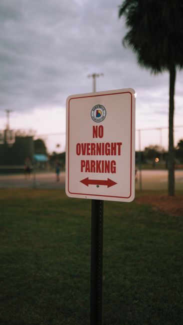 Close-up of a rectangular traffic sign with a white background, red border, and red text reading 'NO OVERNIGHT PARKING' with a red arrow pointing left and right, mounted on a black metal pole situated on a grassy area. In the background, there are tall palm trees, a cloudy sky, and a sports field with a few people and some equipment, indicating an outdoor setting. The scene suggests a location where parking restrictions are enforced to facilitate home relocation or furniture transport processes, as managed by Man with Van Brunswick Park, a professional removals service specializing in house moves and logistics in the Brunswick Park area.
