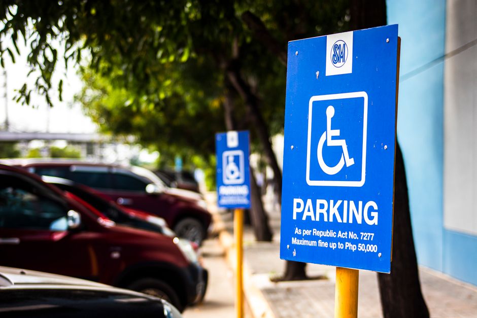A vertical rectangular white 'No Parking' sign with red and black lettering, mounted on a metal pole situated on a street pavement under a partly cloudy sky. In the background, there are no vehicles or people visible. The sign indicates parking restrictions in the area around Brunswick Park, which is relevant for home relocation and furniture transport planning. The image may be used in the context of moving services by Man with Van Brunswick Park, highlighting loading rules and parking regulations necessary to avoid parking fines during loading and unloading processes when conducting house removals or property moves within the area.