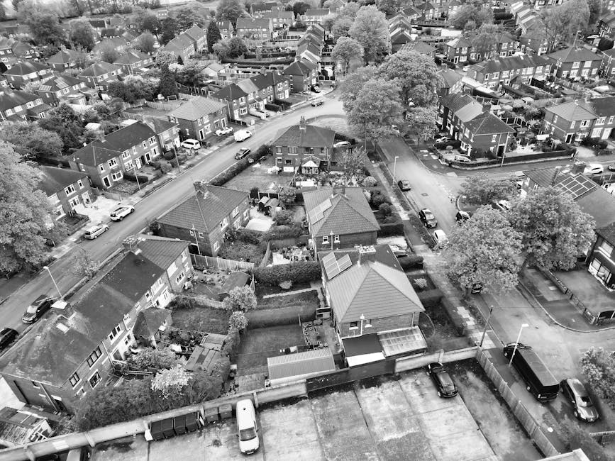 An aerial black-and-white photograph of a residential neighbourhood during a house removal process. The image shows several single-story and two-story houses with pitched roofs, some with solar panels, situated along narrow streets. A large tree with spreading branches is visible in the center, surrounded by gardens enclosed with fences, hedges, and patios. In the foreground, a driveway contains a small white van and various debris or packing materials. The scene captures a manwithvanbrunswickpark.co.uk crew loading or unloading furniture and boxes into a van parked on the pavement, with protective blankets and straps visible on the furniture. The loading area is situated near the entrance to the houses, with some items positioned close to the doorways, indicating a home relocation or furniture transport activity. The overall environment includes parked cars along the street, lamp posts, and neatly maintained gardens, illustrating a typical suburban move coordinated with local street permits in Brunswick Park.