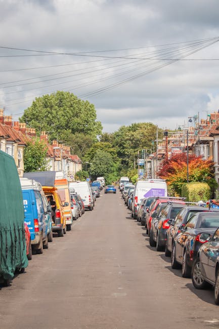 A residential street showing parked cars lining both sides of the narrow tarmac road, with a variety of small terraced and semi-detached houses featuring brick facades and tiled roofs on either side. Some houses have front gardens with bushes and trees, including a prominent green tree at the end of the street and a smaller tree with red leaves on the right. Overhead, there are multiple power lines crossing the cloudy sky. In the foreground, a Man with Van Brunswick Park vehicle is partially visible on the left, covered with a green protective cover, likely used for home relocation or furniture transport. The street appears quiet, suitable for a house removals or moving service, with an emphasis on loading and packing activities typical of household moves. The scene showcases an environment ideal for the logistics involved in local moving services, consistent with the regulations around Brunswick Park and Barnet Council van permits.