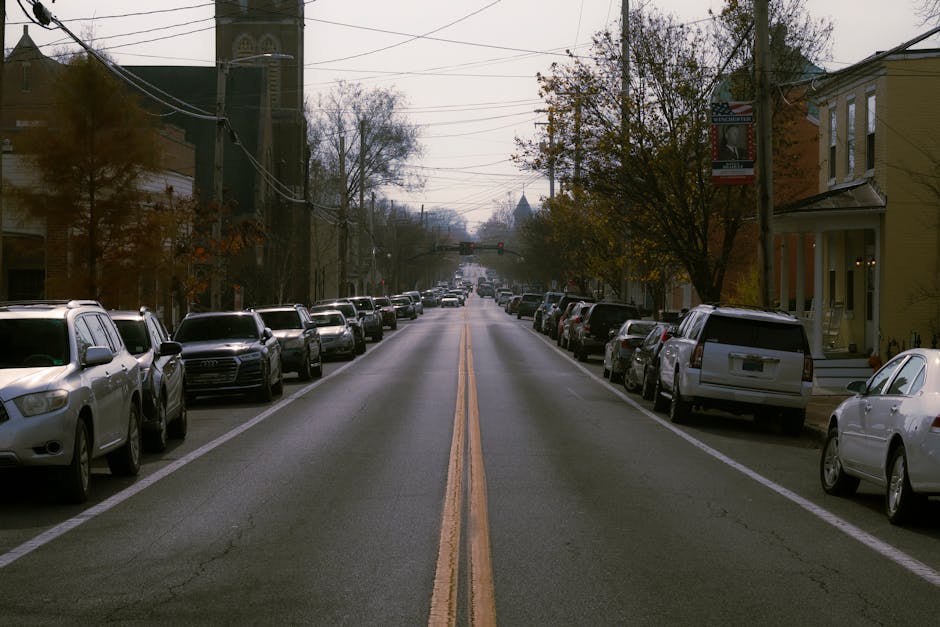 A quiet residential street scene with parked cars lining both sides of the road, including sedans, SUVs, and hatchbacks in various colours such as white, black, and silver. The street has a double yel