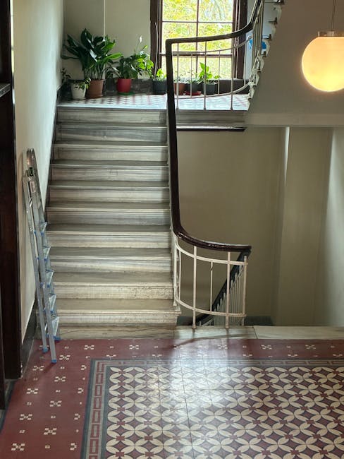 Inside a residential building featuring a staircase with a dark wooden handrail and white balusters, ascending from a patterned tiled floor to a landing with a large window. The window lets in natural light and has several potted plants on its sills, including a tall leafy green plant in a red pot. To the left of the staircase, a blue folding ladder is leaning against the wall, indicating recent or ongoing moving or home renovation activity. The surrounding walls are painted in neutral tones, and a round wall-mounted light fixture provides additional lighting. The image captures the interior environment where furniture and items may be arranged or moved as part of house relocation or furniture transport services. The setting suggests a typical internal stairwell involved in household relocations with attention to safe handling and logistics, as managed by Man with Van Brunswick Park.