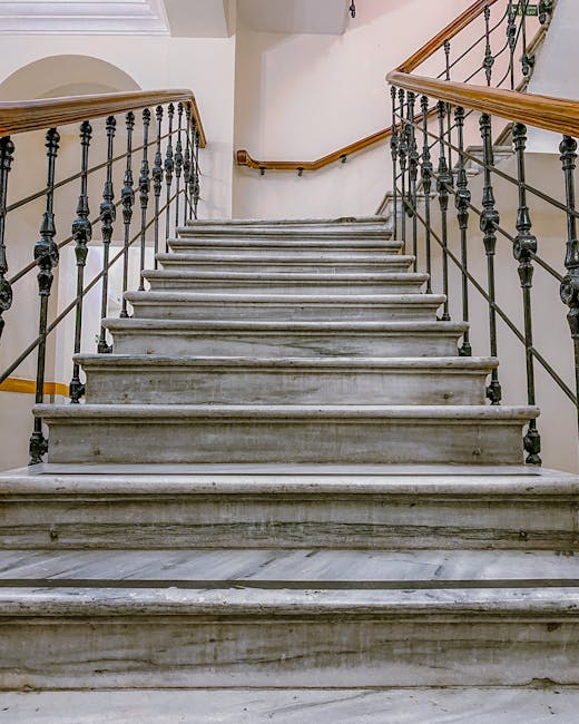 A straight indoor staircase made of grey marble with visible horizontal veining, featuring black metal balusters and wooden handrails on both sides. The staircase is illuminated by natural light coming from an opening or window to the left. The surroundings include a white wall with a partial view of a wooden newel post and a curved wall or archway at the top of the stairs. Inside a residential property, the staircase appears clean and well-maintained, suitable for home relocation or moving activities. This image reflects the interior environment where furniture and bulky items might need to be carefully managed during a house move, with potential considerations for narrow staircases in certain properties. The context suggests a professional removal service like Man with Van Brunswick Park working to transport items through staircases that require detailed packing and careful handling during the loading process.
