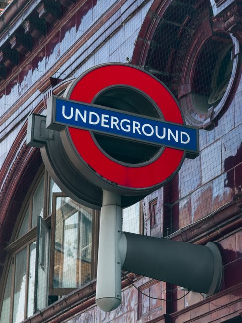 A close-up view of the London Underground roundel sign mounted on a pole outside a building. The sign features a red circle with a blue bar across the middle displaying the word 'UNDERGROUND' in white