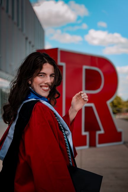A young woman wearing a red academic gown and blue stole stands outside the University of Reading, smiling and holding a small object, possibly a diploma or certificate, in her right hand. Behind her, there is a large red letter 'R' sculpture on a paved area, with a modern building featuring large windows on the left side and a partly cloudy blue sky above. The scene is well-lit with natural daylight, and the woman appears to be celebrating her graduation, with her gown and stole indicating a formal academic event. The image captures the festive atmosphere of a home relocation or moving-related celebration in Brunswick Park as part of the professional services offered by Man with Van Brunswick Park, who handle packing and furniture transport through narrow staircases and complex logistics.