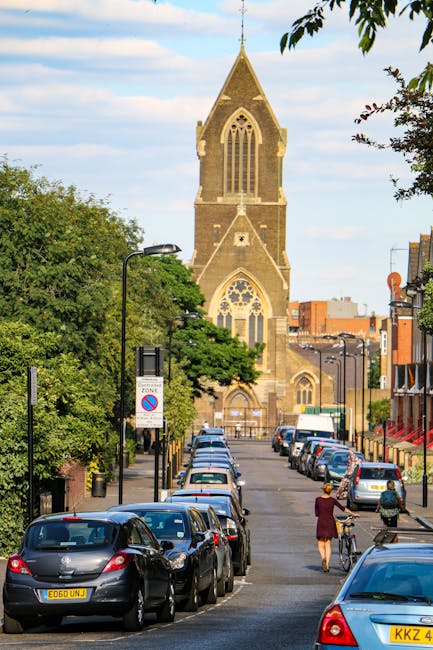 A narrow urban street with parked cars lined along both sides, including hatchbacks, sedans, and small vans, with some vehicles partially occupying the pavement. Two pedestrians are visible—one walking with a bicycle and another nearby—both dressed in casual clothing. Leafy green trees extend along the street, providing partial shade, while a historic stone church with a tall, pointed steeple and large stained glass windows dominates the background. Modern streetlights and traffic signs are present, including a no parking zone sign. The scene is illuminated by natural daylight, suggesting a clear, bright day. This setting captures an active city street suitable for home relocation and furniture transport, reflecting the urban environment in which Man with Van Brunswick Park's removals service operates to facilitate efficient house moving and packing processes.