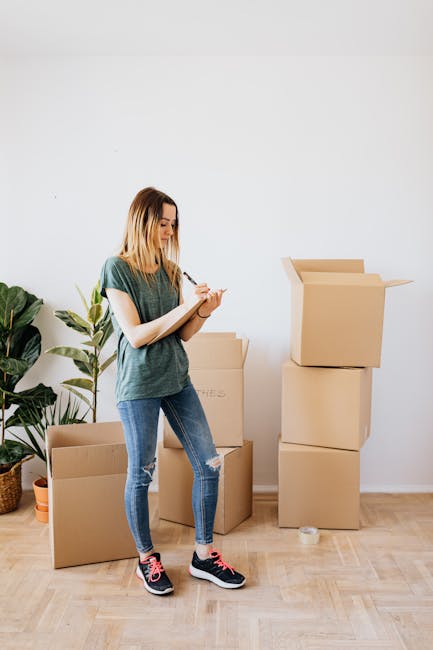 A young woman with shoulder-length blonde hair, dressed in a green T-shirt, ripped blue jeans, and black sneakers with pink accents, stands indoors in front of a plain white wall. She is holding a clipboard and pen, appearing to take inventory or notes related to house removals. To her left, there is a large potted plant with broad green leaves. Behind her, several cardboard moving boxes of varying sizes are arranged; some are stacked, with one box open at the top. The scene suggests an active packing or unpacking process, typical of home relocation, and is set in a clean, well-lit room with a wooden floor. The image is associated with the services of [COMPANY_NAME], a professional removals company, supporting furniture transport, packing, and moving logistics, as part of a house move on the Brunswick Park estate.