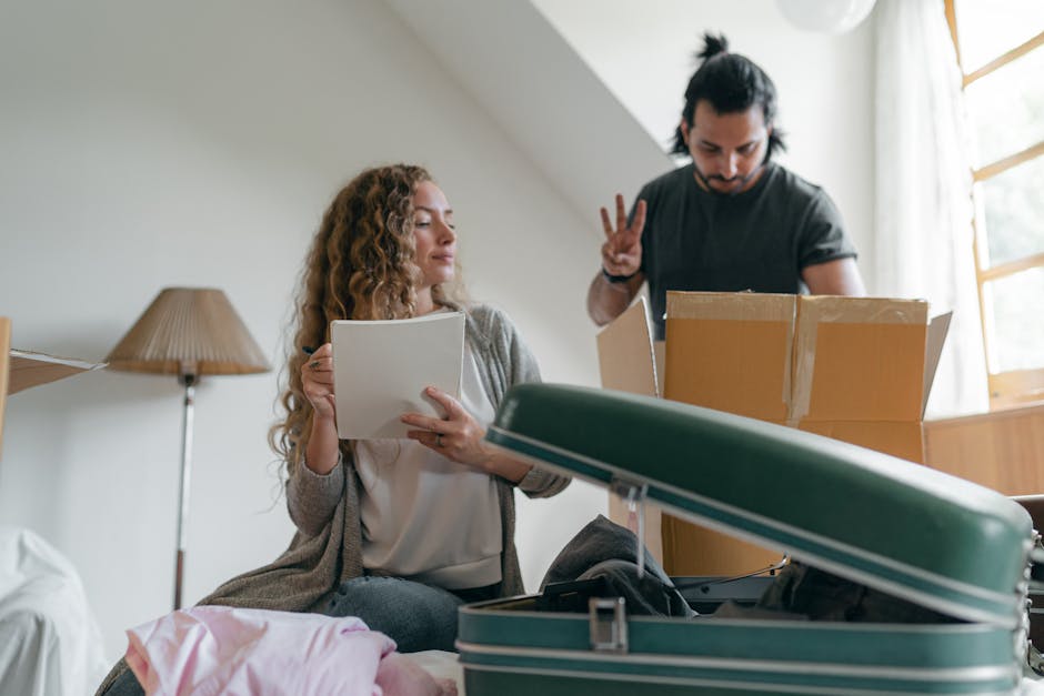 A woman with curly hair, dressed in a beige cardigan and white top, sits on a bed holding a clipboard or folder and appears to be inspecting or reading documents related to house removals. Beside her, a man with dark hair, wearing a black t-shirt, is standing and making a peace sign gesture with his fingers, looking down at the boxes on the bed. In front of them, there is a partially packed, open green and white striped suitcase, along with cardboard boxes, some of which are taped shut. The background shows a well-lit room with a window covered by sheer curtains and a wooden floor lamp. The scene is set indoors, likely in a bedroom or living area, during the packing or home relocation process, with various packing materials and belongings visible, supporting the context of furniture transport and moving services by Man with Van Brunswick Park.