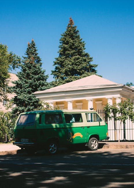A green vintage van parked on the street in front of a white building with a classical portico supported by columns. The van is positioned close to the curb, partially shaded by nearby trees with tall, green foliage. The building behind features a decorative fence, and the scene is set against a clear blue sky, suggesting a sunny day. This scene illustrates a typical home relocation or furniture transport setup, which might be associated with house removals and packing services provided by Man with Van Brunswick Park. The surroundings include paved pavement, and the van's side windows reflect the outdoor environment, emphasizing the vehicular load-in process that could be part of a moving or delivery service.