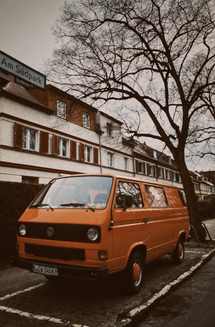 A white moving van parked on a city street in Brunswick Park, with its rear doors open, revealing a variety of packed cardboard boxes, some with visible labels, and plastic bags filled with household items inside. Several cardboard boxes are stacked on top of the vehicle, secured with packing materials, and some boxes are partially open, showing contents. The van is positioned next to a building with multiple stories and large windows, typical of an urban residential area. A small trolley loaded with additional boxes and items is placed on the pavement beside the van, indicating an ongoing packing or loading process. The scene suggests a home relocation or furniture transport operation, assisted by Man with Van Brunswick Park, managing the safe loading of belongings for moving purposes.
