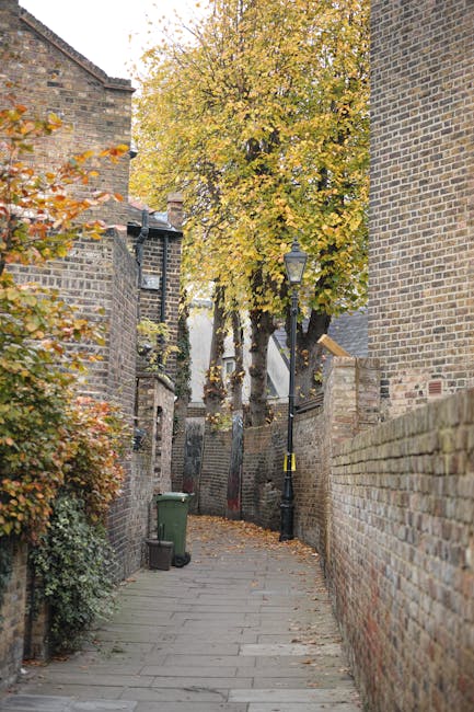 A narrow residential street in Brunswick Park with cobblestone paving, lined with parked cars on the left side and a building façade on the right. The building features black wrought-iron lanterns at