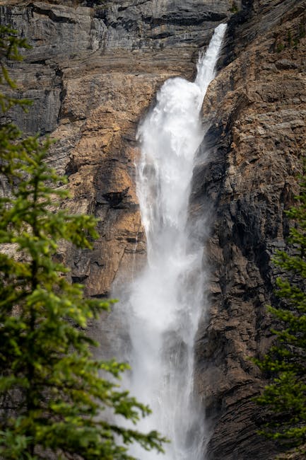 A tall waterfall cascading down a rugged, rocky cliff face with water forcefully flowing over the edge, creating white spray and mist. Surrounding the waterfall are green trees and foliage, some visible at the bottom and sides of the image. The scene is set outdoors in a natural environment with bright, natural lighting highlighting the texture of the rocks and the movement of the water. The image captures the power and height of the waterfall, emphasizing its presence within a scenic landscape. This natural scenery can be associated with the concept of moving or transporting items through outdoor environments, aligning subtly with house removals and logistics services offered by companies like Man with Van Brunswick Park.